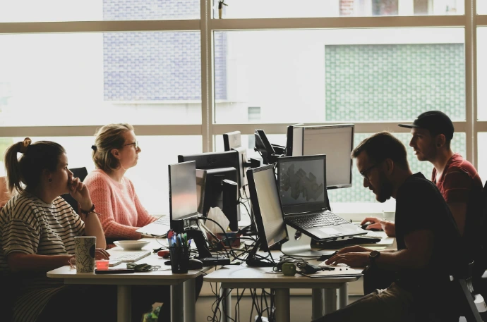 Four people working at computers in a sunlit office workspace